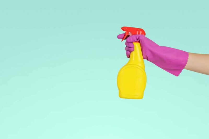 A pink-gloved hand holds a yellow spray bottle in front of a turquoise background