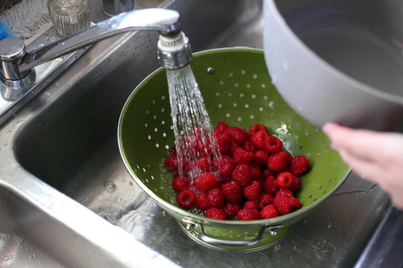 A person washing a bowl of raspberries in a sink.